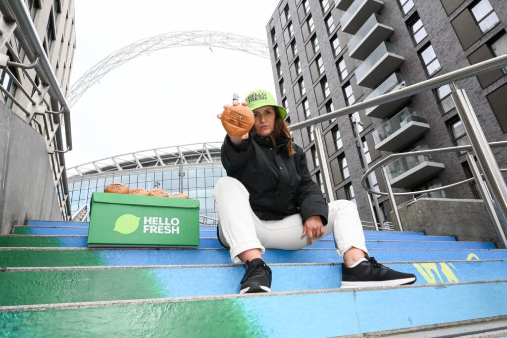 A HelloFresh courier in front of Wembley Stadium holds a bread roll with "roll with it" printed on it.