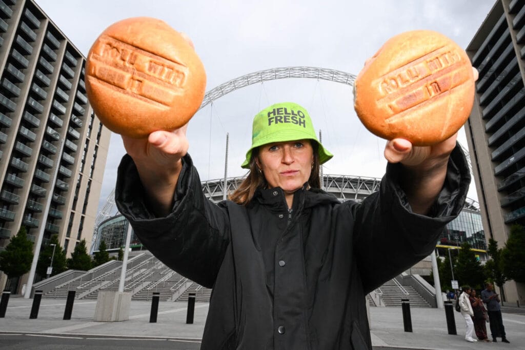 A HelloFresh courier stands in front of Wembley Stadium holding two bread rolls with "roll with it" printed on.